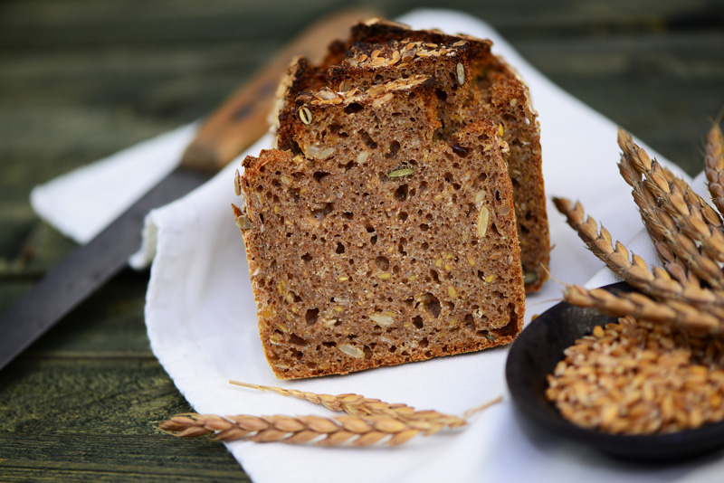 Ein angeschnittenes Vollkornbrot liegt auf einem Tuch, man sieht die volle Bandbreite an Körnern und Samen, davor steht eine Schüssel mit Getreidekörnern und daneben liegt eine Ähre vom Feld. Im Hintergrund sieht man noch leicht das Messer.