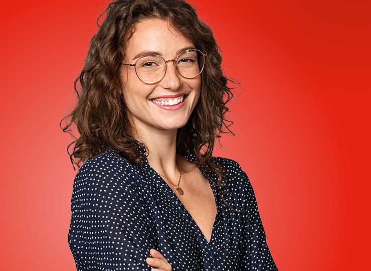 Studentin mit braunen schulterlangen Locken und Brille schaut breit lächeln. Sie trägt eine dunkelblaue Bluse mit weißen Punkten und V-Ausschnitt. Sie hält die Arme vor der Brust leicht verschränkt.
