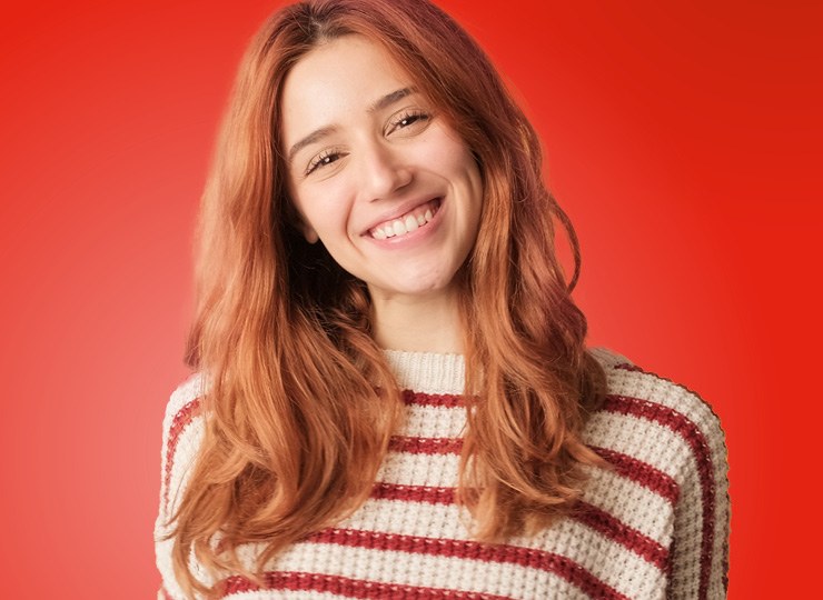 Junge Frau mit roten, langen Haaren lächelt breit. Sie trägt einen rot-beige-gestreiften Pullover.