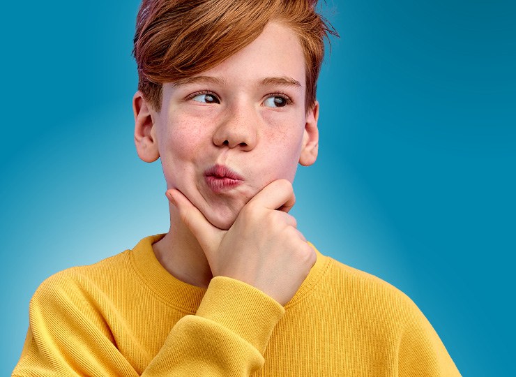 Junge mit roten kurzen Haaren trägt einen gelben Pullover. Er hat die hnd am Kinn, verzieht den Mund und überlegt.